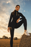 Woman in black athletic wear with a logo, standing on a hay bale in a field.