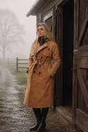 Woman in a tan trench coat standing in front of a wooden shed on a foggy day.
